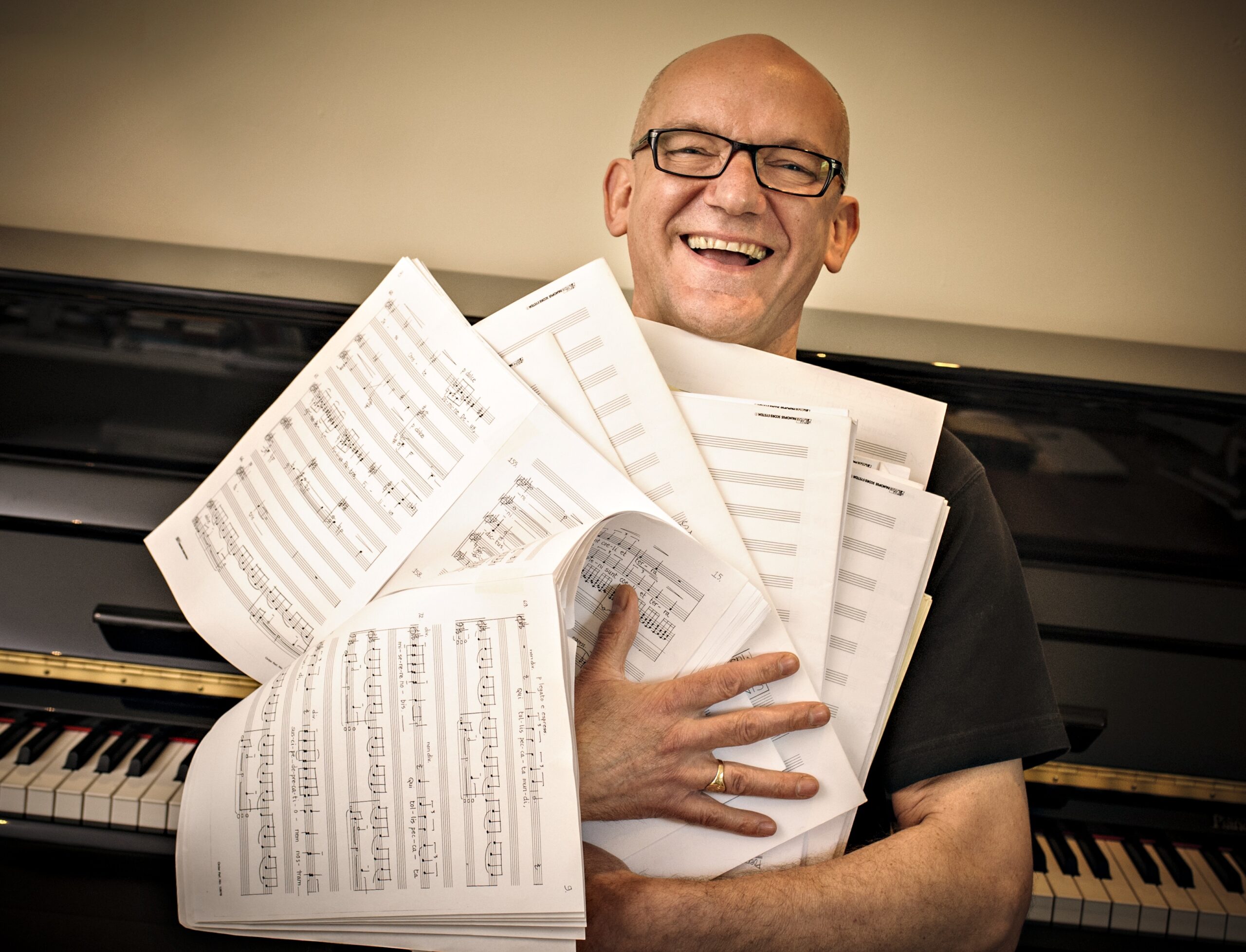 Broadly smiling bald man wearing dark-rimmed glasses, with his arms full of a tumble of sheets of music, like he's hugging them. Black piano, with keys showing, and cream wall behind him. (Composer Bob Chilcott)