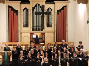 About 40 women singers from Durango Women's Choir and SF Women's Ensemble, all wearing black and standing on steps. Large pipes of an organ in the background (tan, dull metal, and brown wood) (2015)