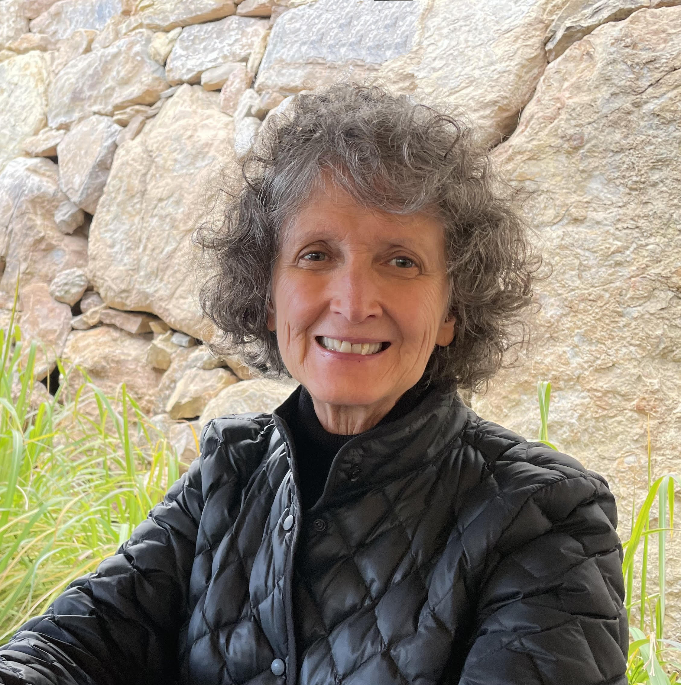 Smiling woman with short curly dark grey hair wearing black quilted jacket, sitting in front of tan stone wall and green grass below (Director Linda Raney)