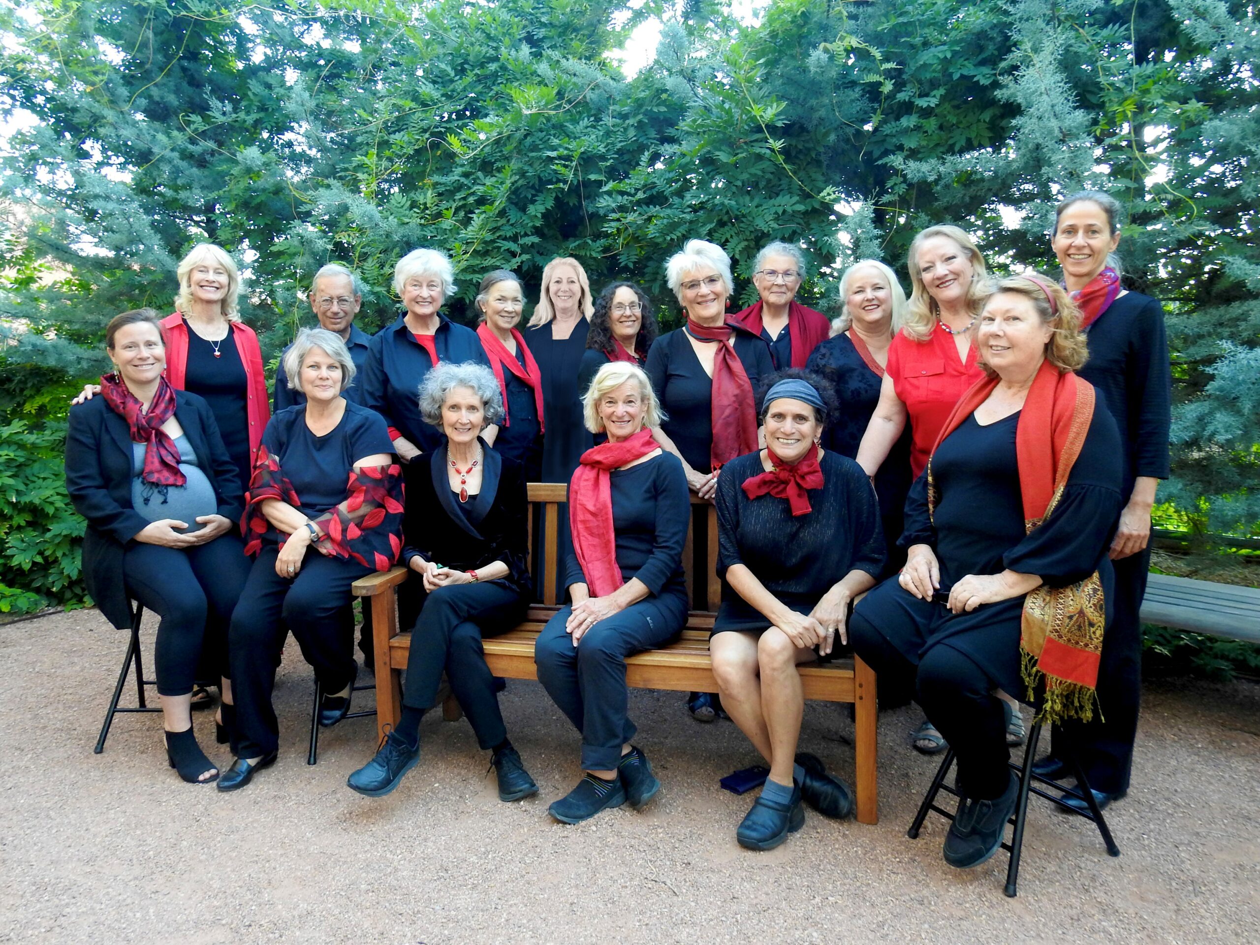 Group of smiling women (the singers and director) both seated and standing. One man, the accompanist. All wearing black, some with red scarves, red shawls or red shirts. Seated on bench and stools, gray sand underfoot and bright greenery as the background.