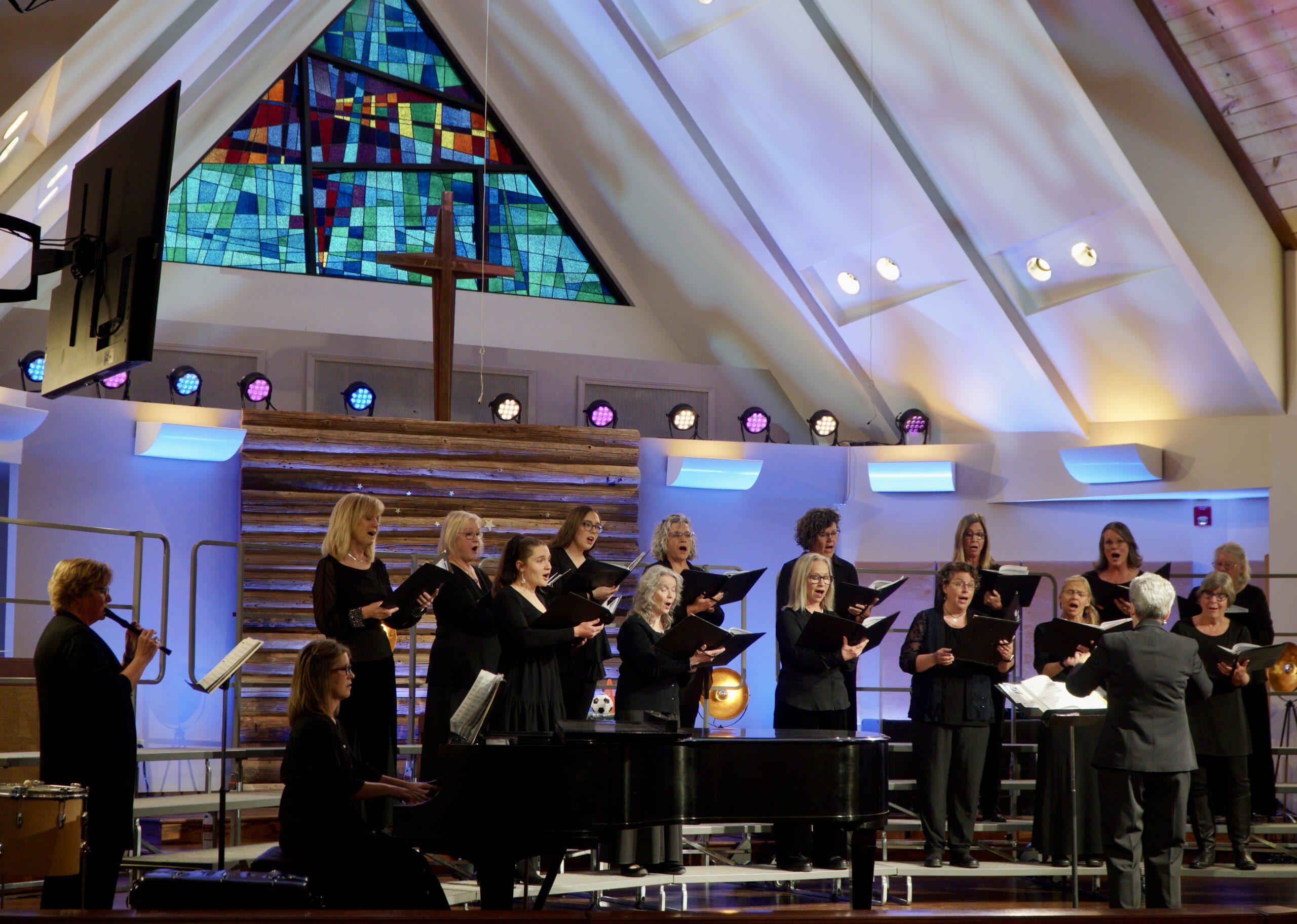 Small group of women wearing black singing while standing on risers. Background has A-frame ceiling with abstract stained glass window which creates blue and lavendar shadows on  the walls. 