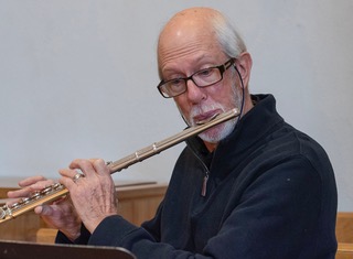 Man with fringe of grey hair and a beard (wearing black) playing flute