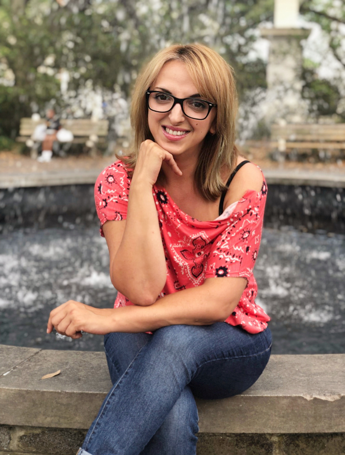 Smiling woman with shoulder-length light brown hair, wearing dark glasses, pink/red shirt and blue jeans, sitting by fountain (blurry trees in background)