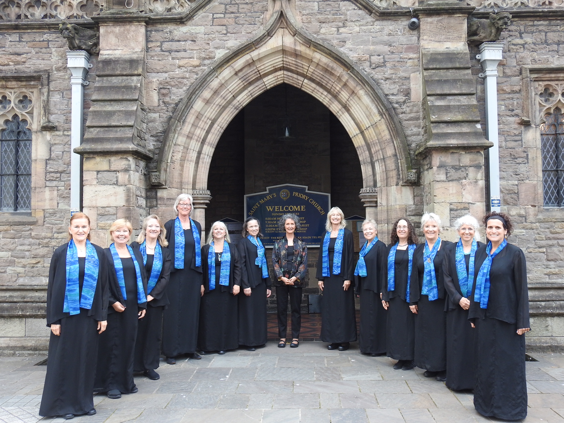 Group of singers, wearing black with turquoise scarves, standing in front of stone church entrance, Abergavenny, Wales, 2018