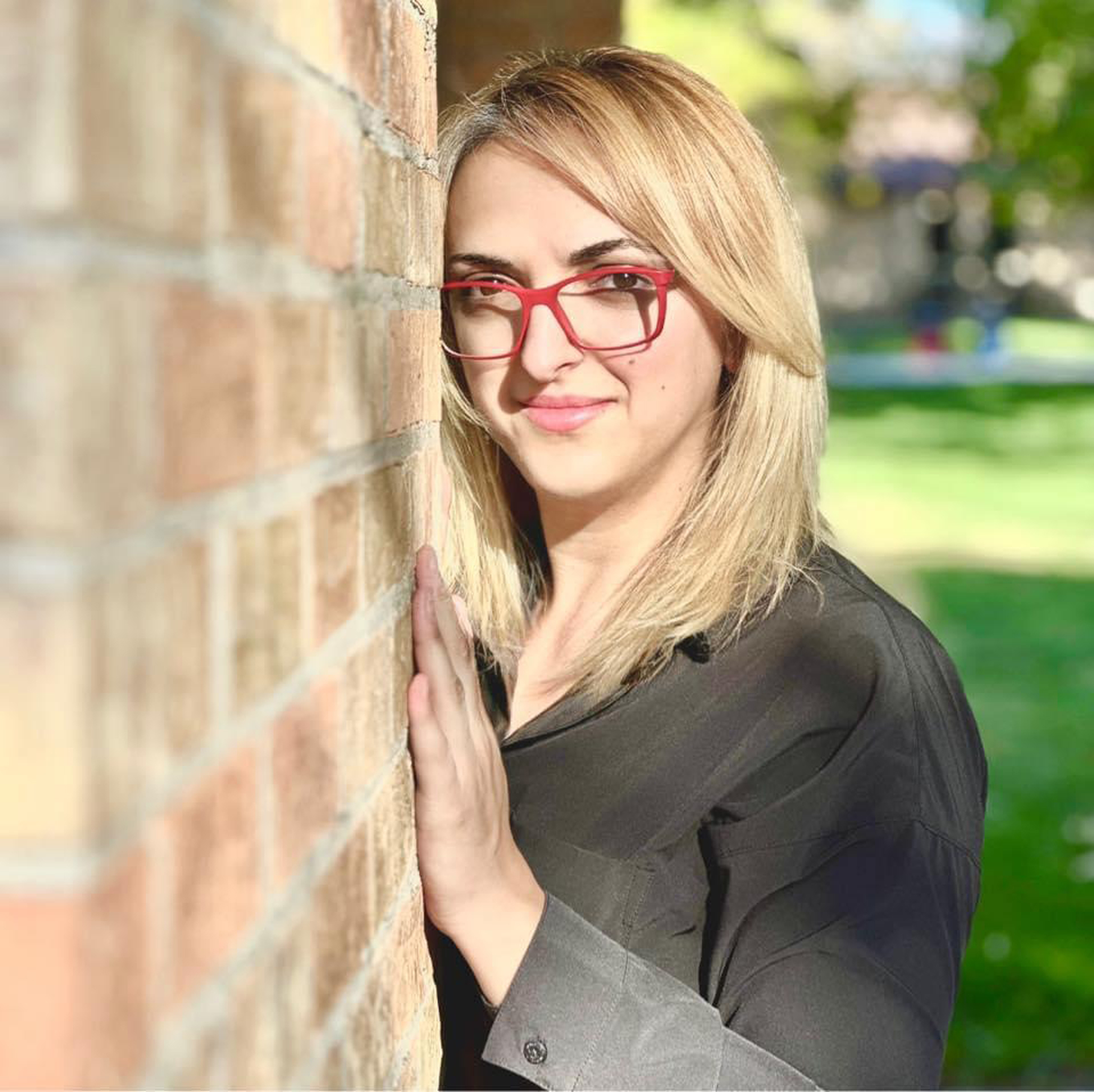 Woman with blonde hair with red glasses and wearing black, leaning with hand against brick wall