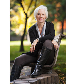 Woman with short silver hair, sitting on the arm of a bench in a park, leaning with arms crossed on her knee