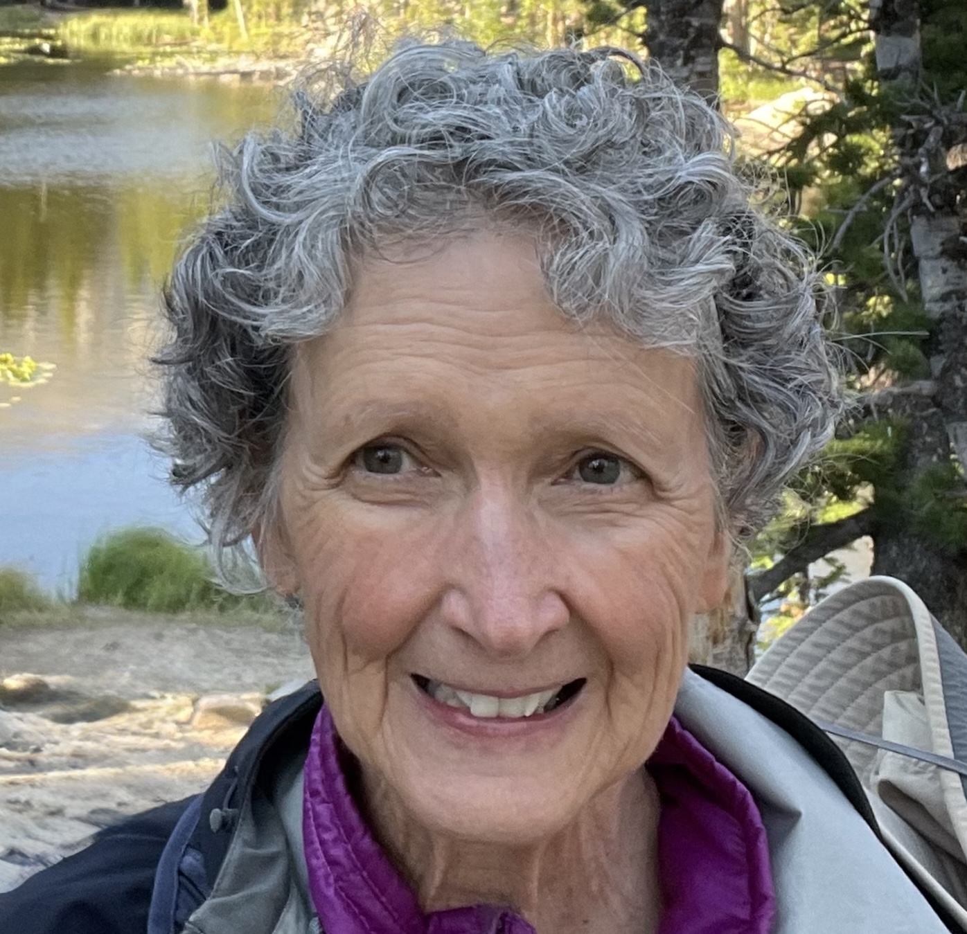 Linda 2025 Linda Raney, smiling with short tousled grey curls. Serene lake behind her.