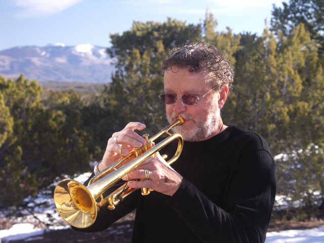 Man playing shiny golden trumpet, background of piñon trees and distant mountain