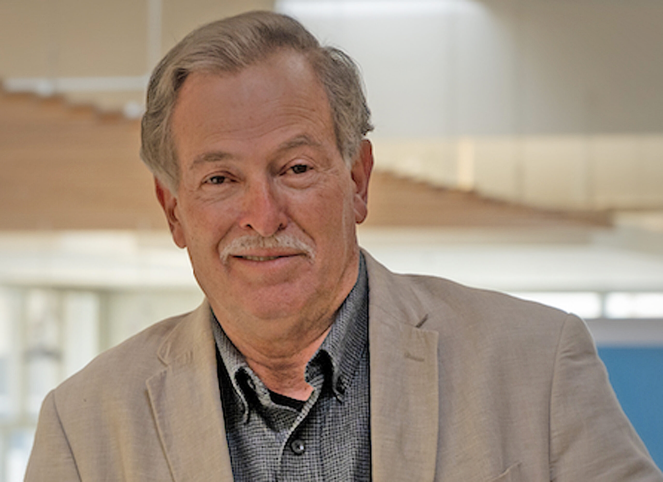 Smiling man with grey hair and mustache wearing blue shirt and tan jacket
